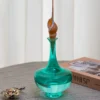 Bouilloire en verre vert avec bouchon décoratif en coquillage, placée sur une table gris clair.