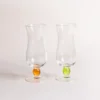 Deux verre à eau transparents avec pied perlé en verre orange et vert pomme, posés sur une table claire sous une lumière douce.