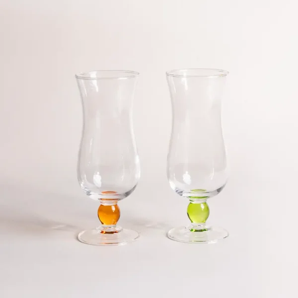Deux verre à eau transparents avec pied perlé en verre orange et vert pomme, posés sur une table claire sous une lumière douce.