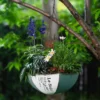 Joli pot de fleur rose à suspendre, contrastant avec les fleurs jaunes et violettes dans un jardin luxuriant.