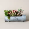 Pot de fleurs moderne en métal galvanisé avec plantes grasses, orné du logo “NEST” pour une décoration contemporaine.