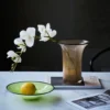 Vase pour tulipes minimaliste posé sur une table ensoleillée avec fruits et orchidées.