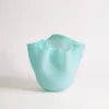 Vase art déco turquoise en verre soufflé, mis en valeur avec des fleurs blanches et une lumière naturelle douce sur fond de stores.